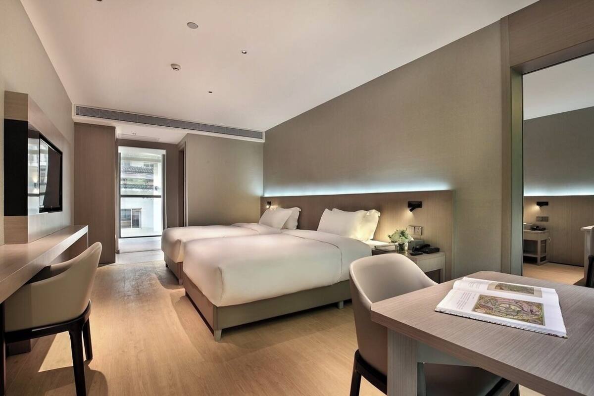A modern hotel room with a large bed, a desk, and a chair. There is a window providing a view of the outside.