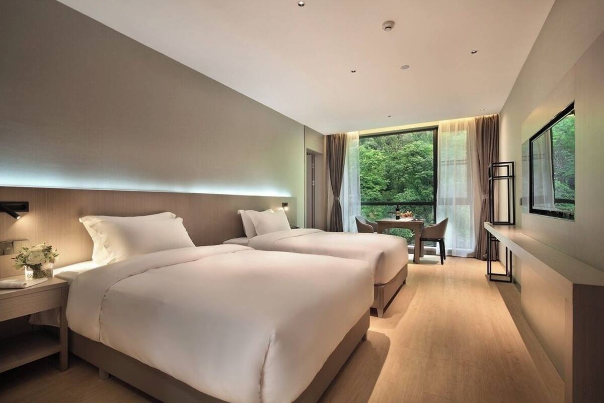 A modern hotel room with two beds, a dining area, and a large window offering a view of the outdoors.