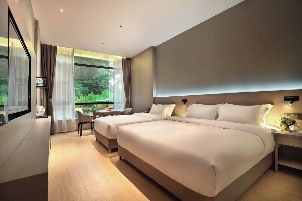 A modern hotel room with a large bed, a desk, and a view of greenery through the windows.