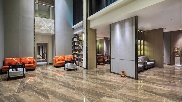 Een ruime lobby met een hoge plafond, marmeren vloer en moderne meubels, waaronder oranje en grijze banken.