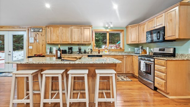 A spacious kitchen with wooden cabinets, a large island with bar stools, stainless steel appliances, and a view of the outdoors. - Cozy 3-bedroom cabin in charming Haverhill with Hot Tub, Play Area & Fire Pit (Haverhill)