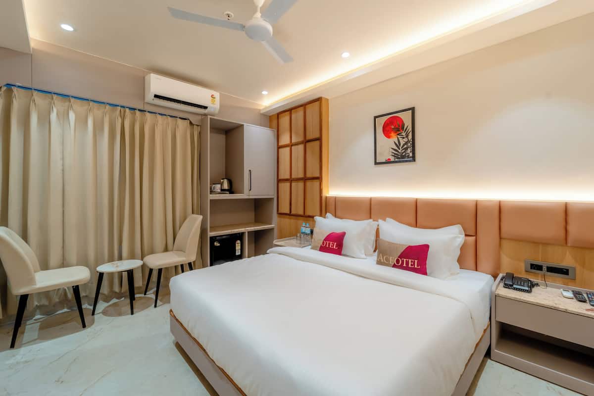 Quarto de hotel com cama, duas cadeiras, escrivaninha, guarda-roupa e ventoinha no teto.