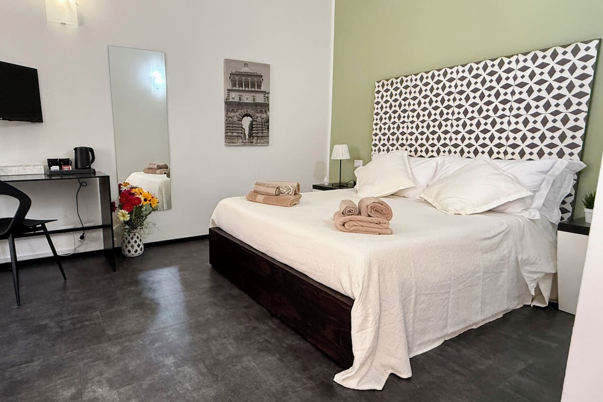 A hotel room with a bed, a desk, a chair, a TV, a mirror, and a wall with a geometric pattern.