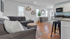 A modern living room with a grey sofa, a round ottoman, and a kitchen area with white cabinets and a black stove.