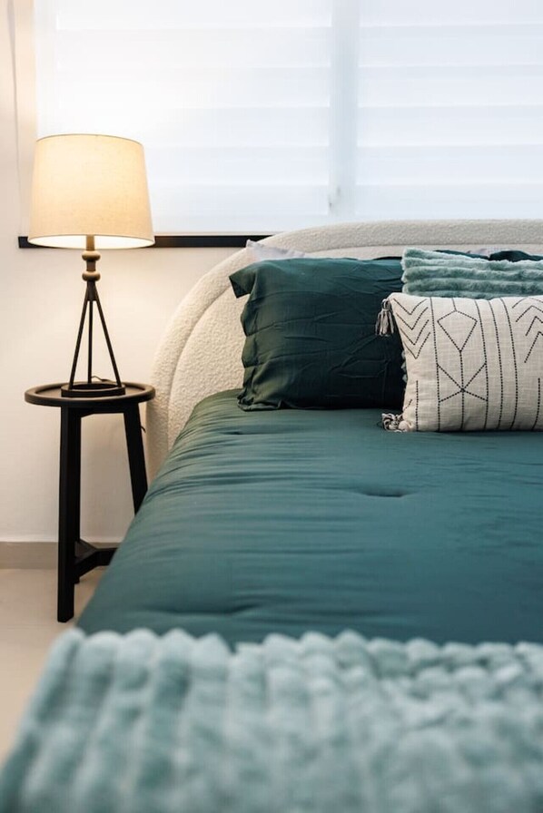 A neatly made bed with a green bedspread and pillows, a small black side table with a lamp, and a window with blinds in the background. - Modern & Spacious Retreat in Hato Rey | Unit 2 (San Juan)