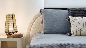 A neatly made bed with a grey and white color scheme, a bedside lamp with a geometric pattern, and a window with horizontal blinds. - Modern & Spacious Retreat in Hato Rey | Unit 2 (San Juan)