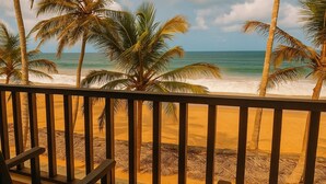 Une vue sur la plage depuis un balcon doté de balustrades en bois, avec des palmiers et les vagues de l’océan en toile de fond.