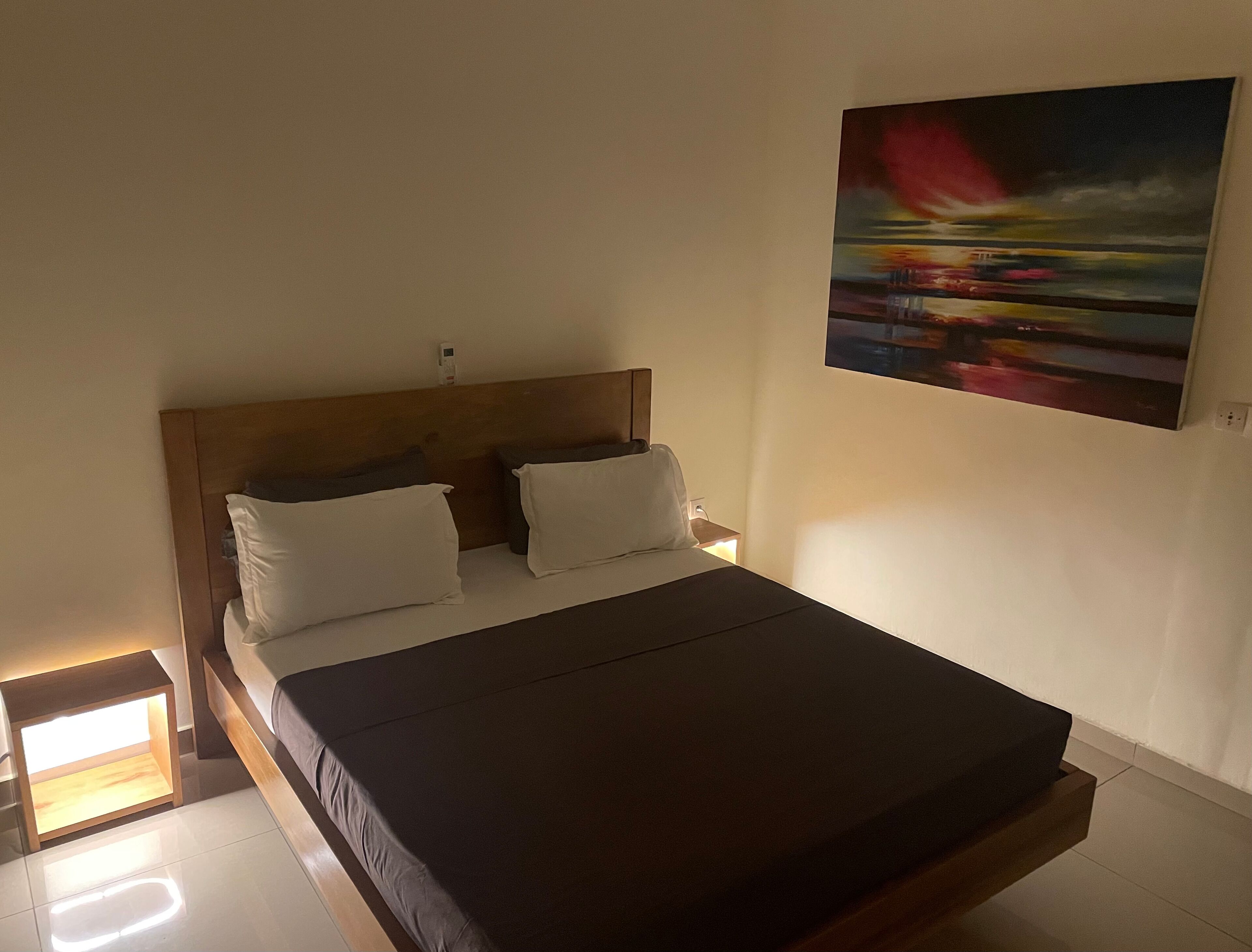 Un lit simple avec une tête de lit en bois, des oreillers blancs et une couvre-lit sombre, dans une chambre équipée d’une table de chevet et d’un tableau accroché au mur.