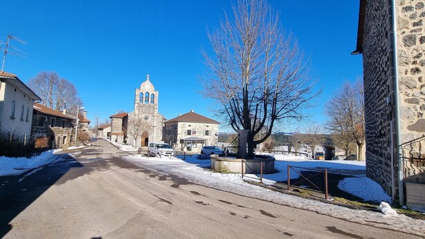 A snow-covered street with stone buildings, a church with a bell tower, and parked cars.