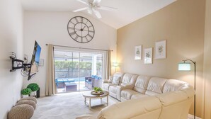 A living room with a sofa, a coffee table, a ceiling fan, a clock, and a view to the outside.