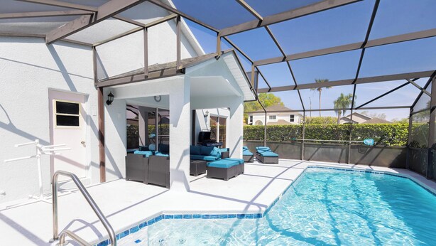 A swimming pool with a metal ladder, a covered seating area with blue cushions, and a white exterior wall.