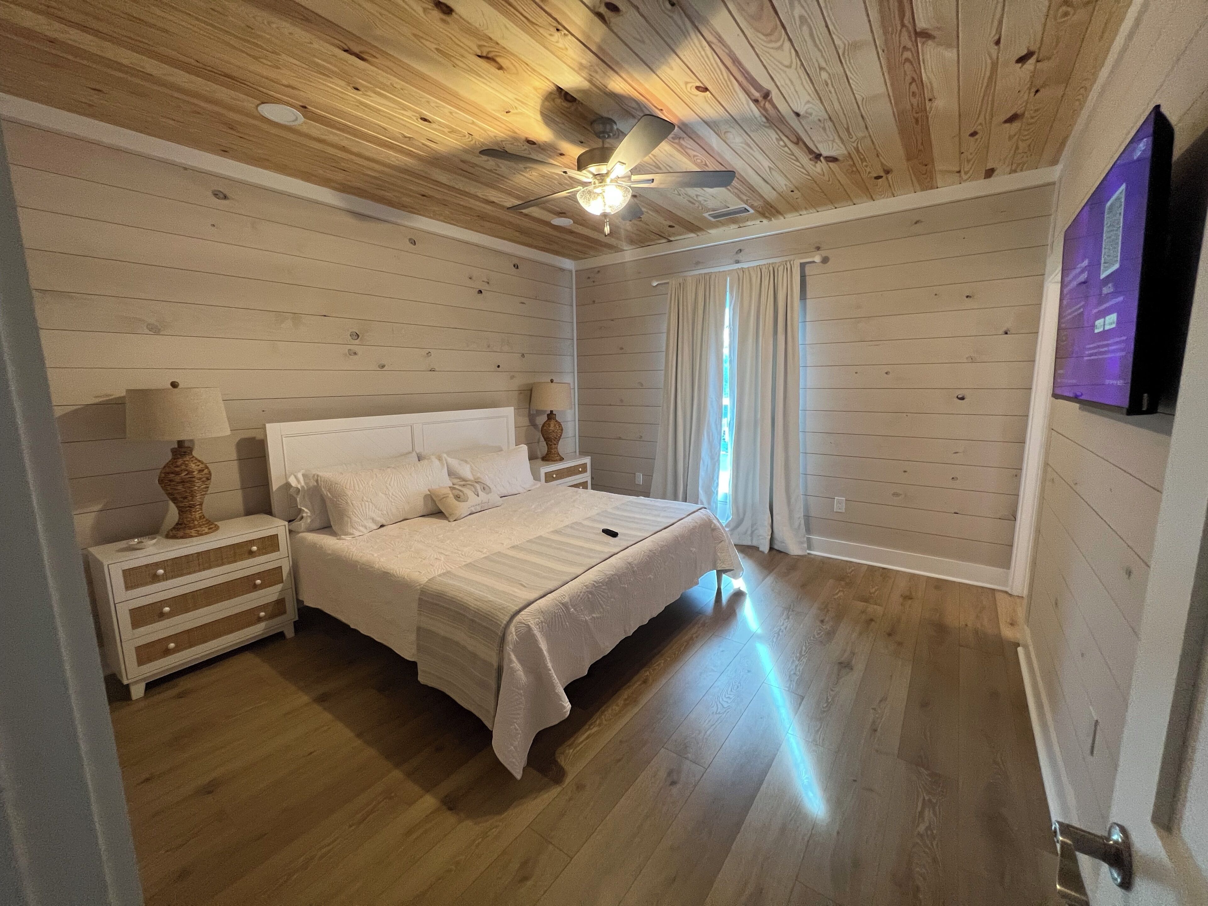Une chambre avec un plafond en bois, un lit recouvert de linge de lit blanc, deux tables de chevet avec des lampes et un téléviseur fixé au mur.
