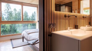 A modern bedroom with a large window, wooden paneling, a bed, a bedside table, and a bathroom with a sink and mirror. - Chalet Eteila Combloux proche Megève (Combloux)