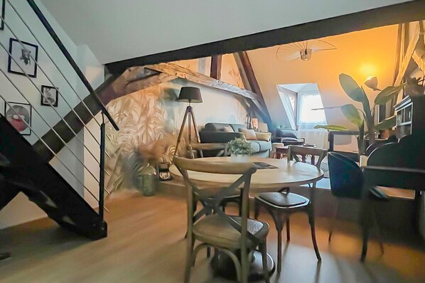 A modern living room with a sofa, dining table, and chairs. There is a staircase leading to an upper level.