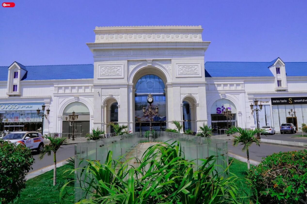 A white building with a central arched entrance, surrounded by a glass atrium and palm trees.