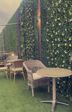 Uma área externa com assentos de vime, cadeiras e uma mesa redonda, cercada por um muro verde e flores brancas.
