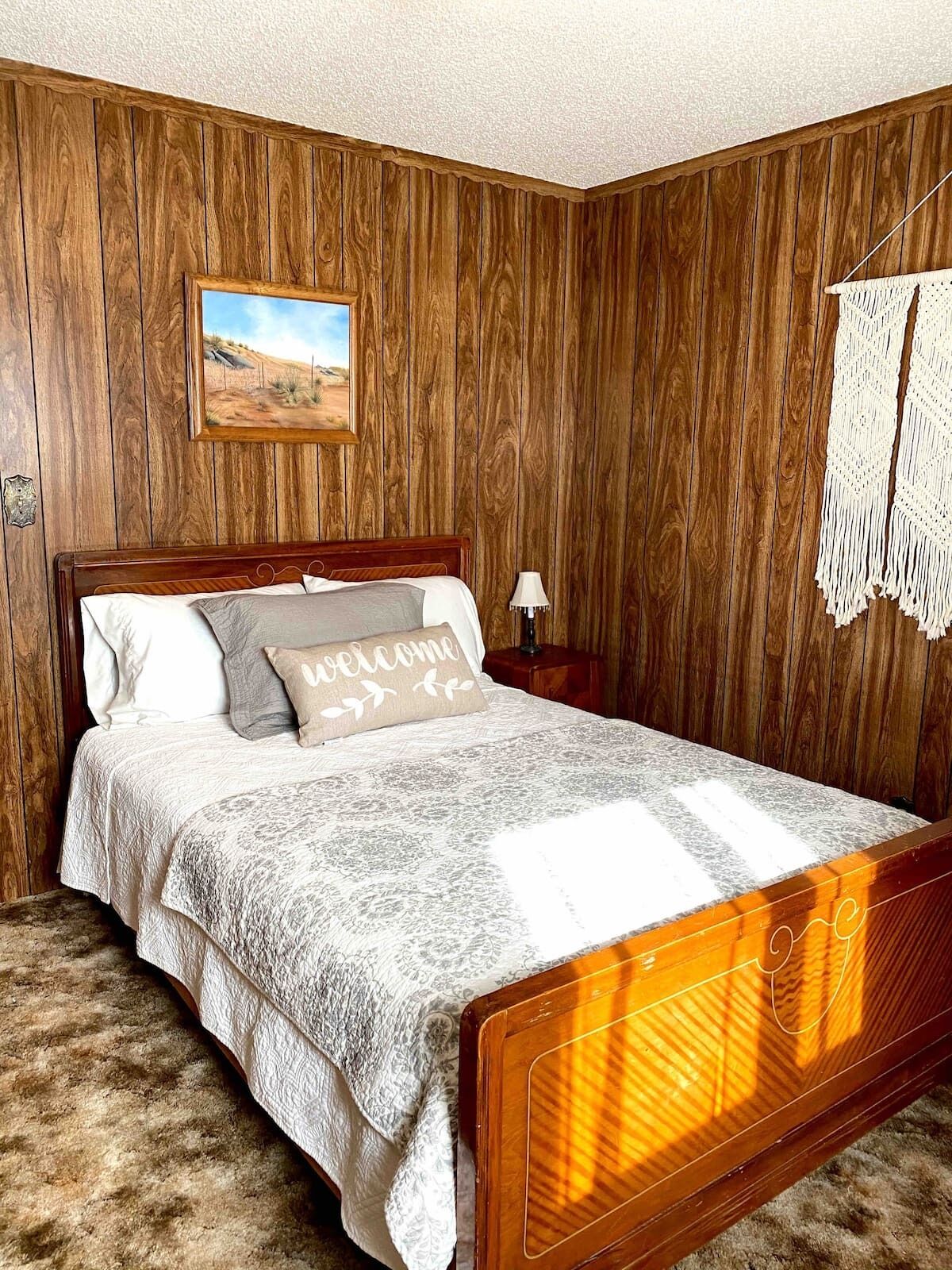Un dormitorio con una pared revestida de madera, una cama con colcha blanca y gris estampada, una almohada de bienvenida y un cuadro enmarcado sobre la cama.