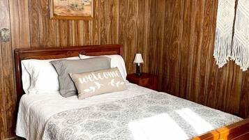 Un dormitorio con una pared revestida de madera, una cama con colcha blanca y gris estampada, una almohada de bienvenida y un cuadro enmarcado sobre la cama.