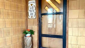 Fachada de madera con puerta de vidrio de marco negro, un cartel de bienvenida y una maceta pequeña con flores.