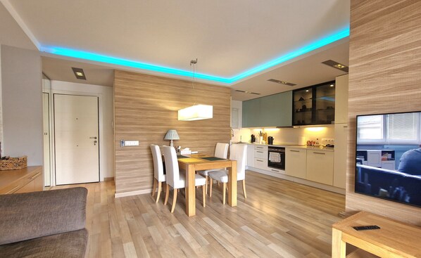 A modern living room with a dining area, wooden flooring, a flat-screen TV, and a kitchen with built-in appliances. - Apartment Columbo Go2lloret (Lloret de Mar)
