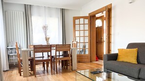 Ein Wohnzimmer mit einem Glastisch, Holzstühlen, einer gelben Kissen ausgelegten Couch und einem Essbereich mit Tisch und Stühlen.