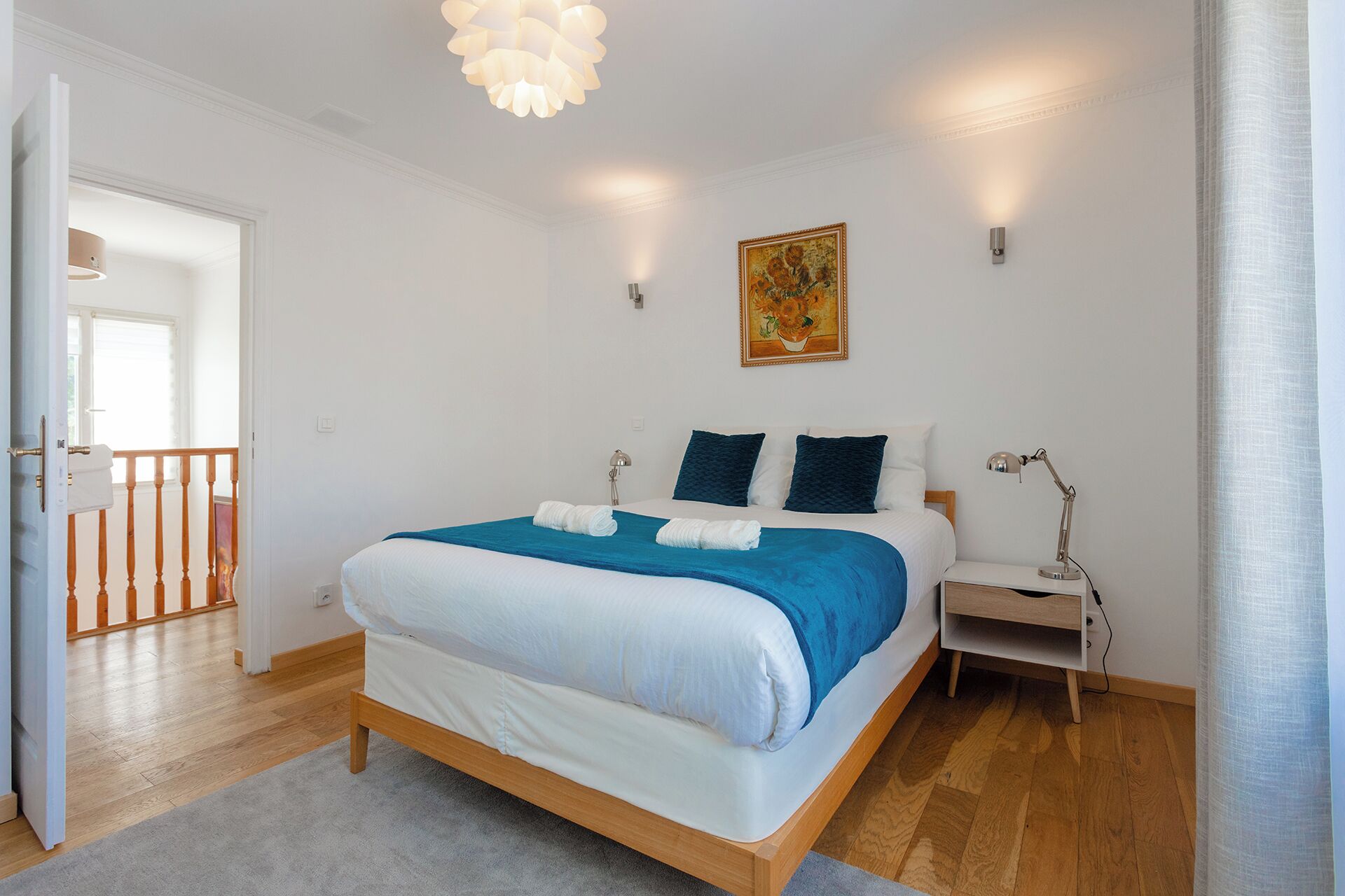 Ein Schlafzimmer mit einem hölzernen Bett, weißer Bettwäsche, einem blauen Akzent, einem Nachttisch, einer Schreibtischlampe und einem Gemälde an der Wand.