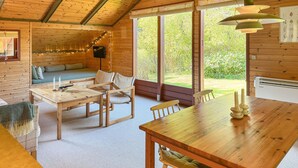 L’intérieur chaleureux d’un chalet avec une table à manger en bois, des chaises, un canapé et de grandes fenêtres donnant sur un paysage verdoyant.