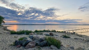 Une plage avec un rivage sablonneux, des rochers et une rambarde métallique surplombant une étendue d’eau calme.