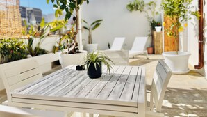 En terrasse med et hvidt bord og stole, omgivet af krukker med planter og en klar blå himmel.