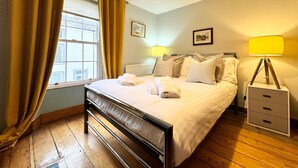 A neatly made bed with pillows and towels, a bedside lamp, a nightstand, and a window with curtains. - The Cottage, 3 Bedroom, Pets Welcome (Ulverston)