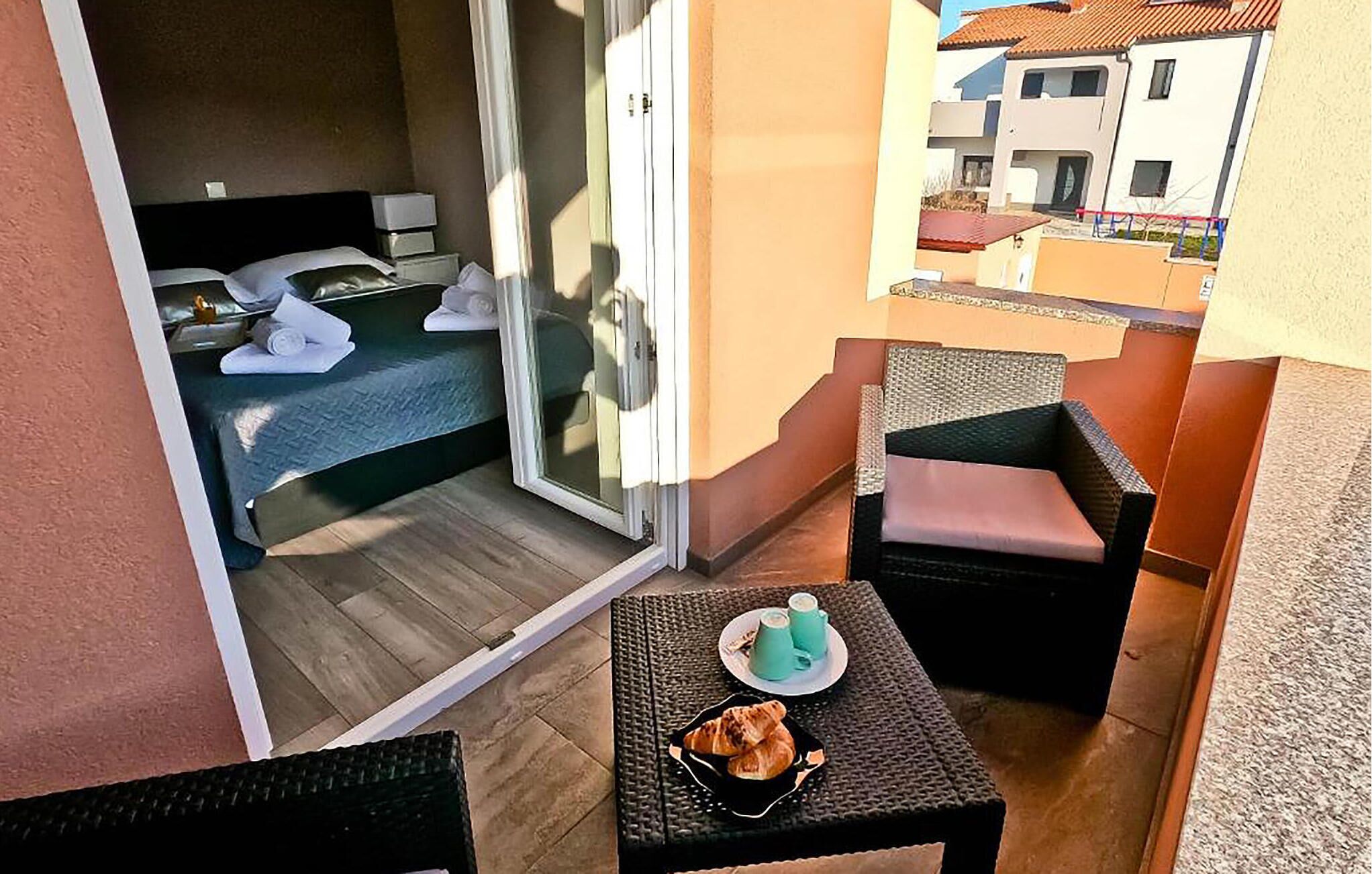 A balcony with a bed, two chairs, a small table with a plate of croissants and two cups, and a view of buildings.