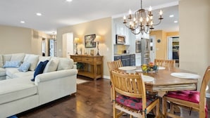 A spacious living room with a sofa, dining table, and chairs. There is a kitchen in the background with cabinets and appliances.