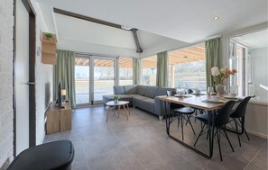 A modern living room with a sofa, dining table, and chairs. There is a large window with curtains, a small table with a plant, and a TV on a stand. - Gorgeous home in Stavenisse (Stavenisse)