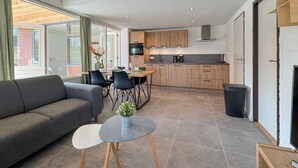A modern kitchen with wooden cabinets, a dining area with a grey sofa and chairs, and a small table with a plant.
