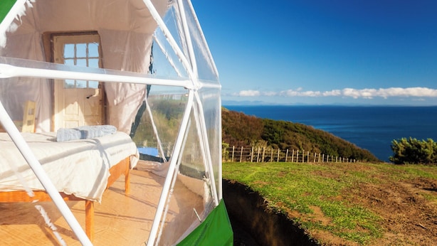 A geodesic dome structure with a bed inside, overlooking a coastal landscape with a clear sky. - Escarpment Domes (Porirua)