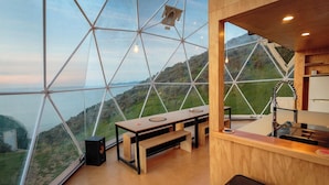 A modern room with a large glass window offering a panoramic view of a green landscape. There is a wooden table with two chairs, a small stool, and a kitchen area with a sink and countertop. - Escarpment Domes (Porirua)