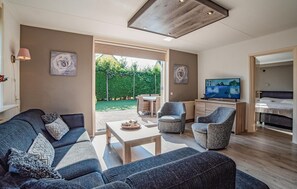 A modern living room with a sofa, armchairs, a coffee table, and a TV. There is a garden visible through the glass door.