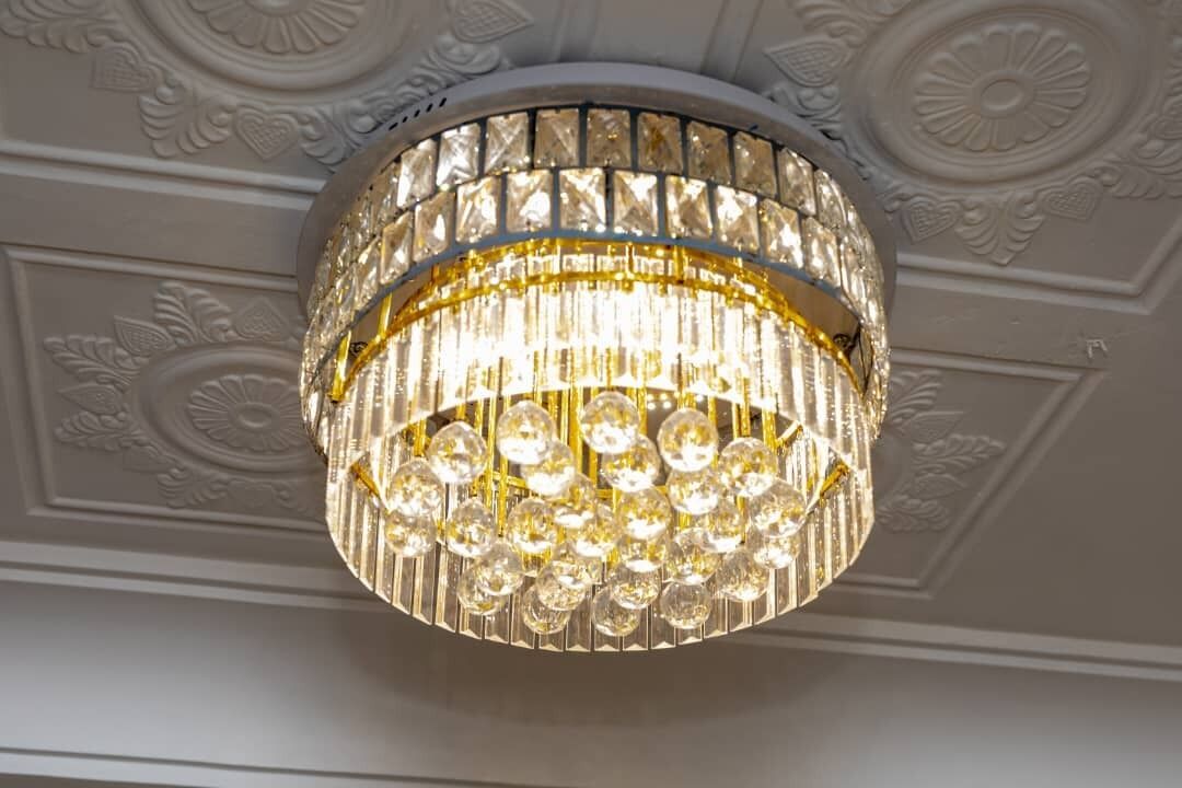 A chandelier with multiple clear crystal elements and gold accents, set against a decorative ceiling with embossed floral patterns.