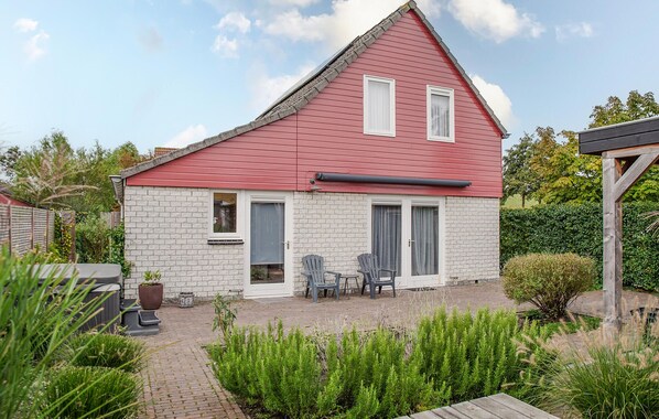 Two-story house with a red roof and white brick walls, featuring two windows with white shutters and a front door with a glass panel, set amidst a garden with lush greenery.