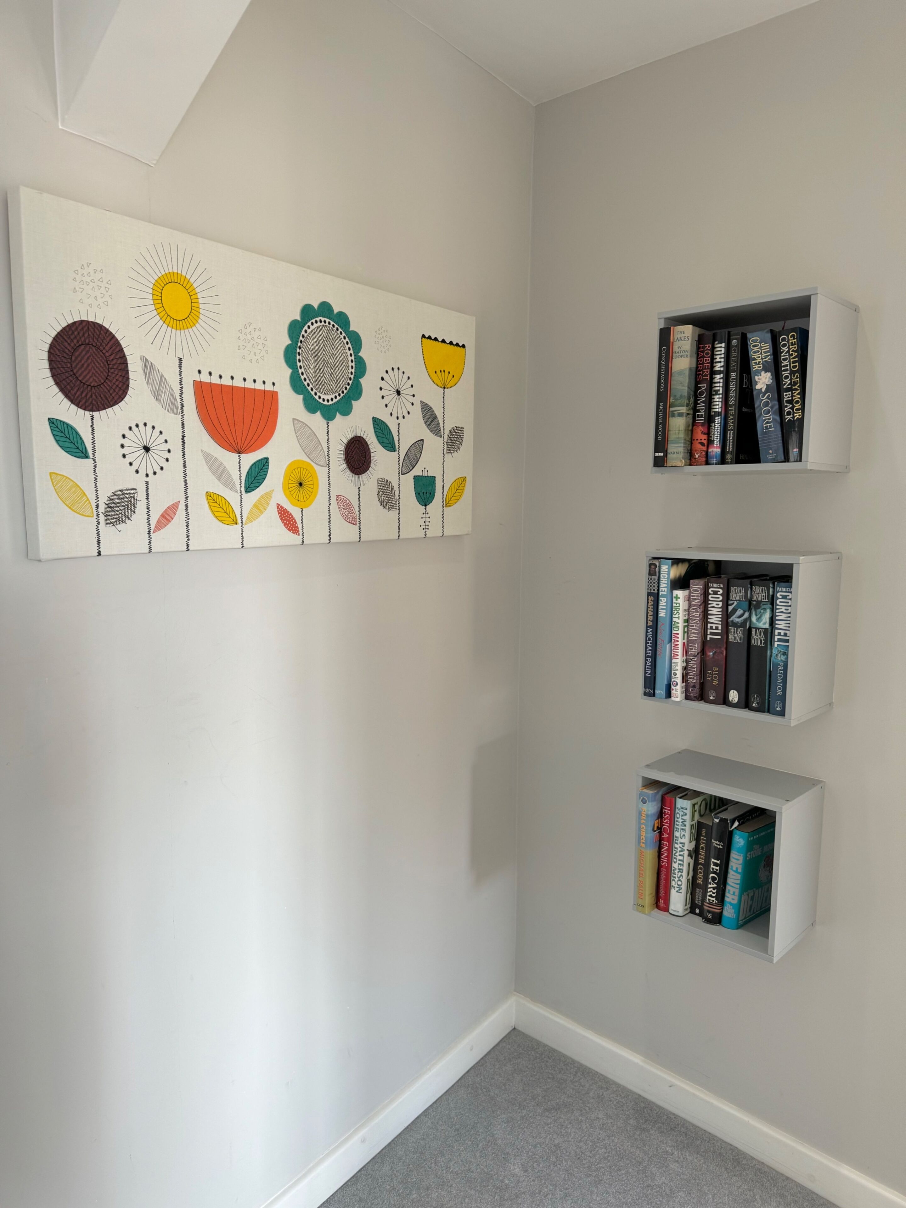 A room with a white wall, a colorful floral artwork, and three white bookshelves filled with books.