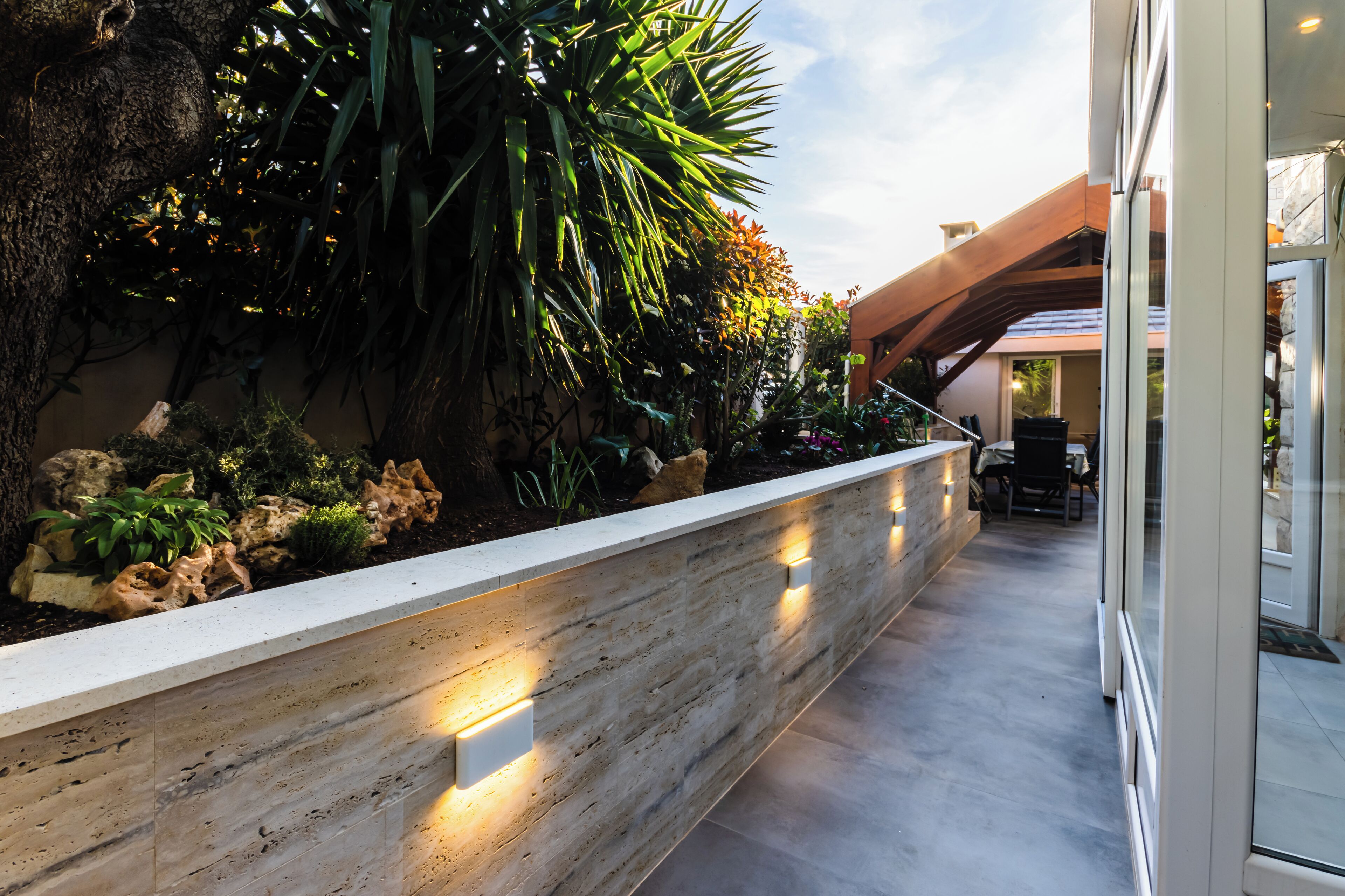 A modern outdoor area with a stone planter, various plants, and a seating area with a table and chairs.