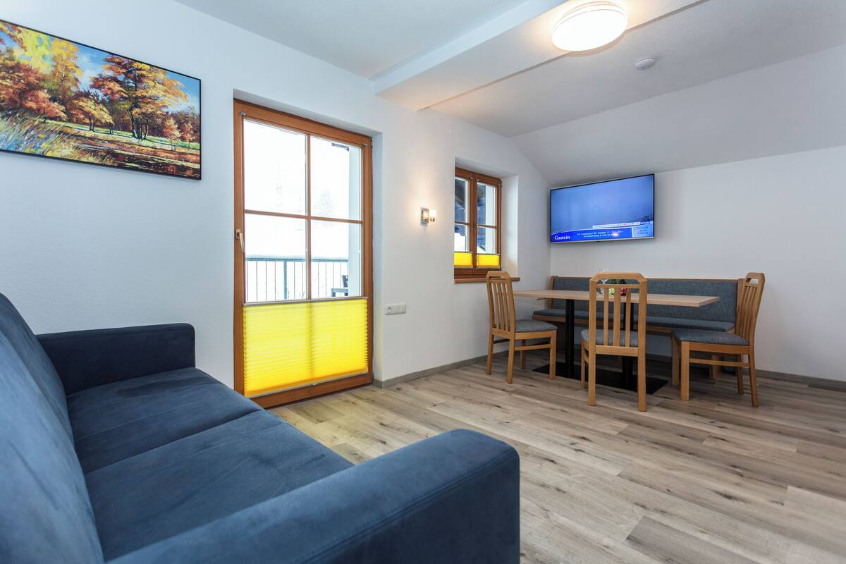 A living room with a blue sofa, a dining table with chairs, a TV mounted on the wall, and a window with blinds.
