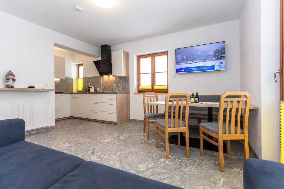 A modern kitchen with white cabinets, a built-in oven, and a stone countertop. There is a dining area with a wooden table and chairs, and a TV mounted on the wall.