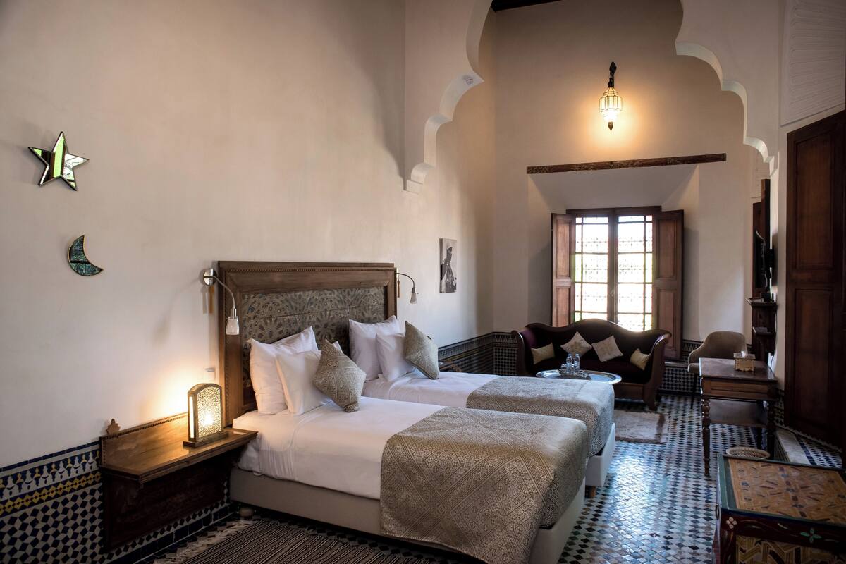 A room with a bed, two bedside tables, a nightstand, a chair, and a window with natural light.