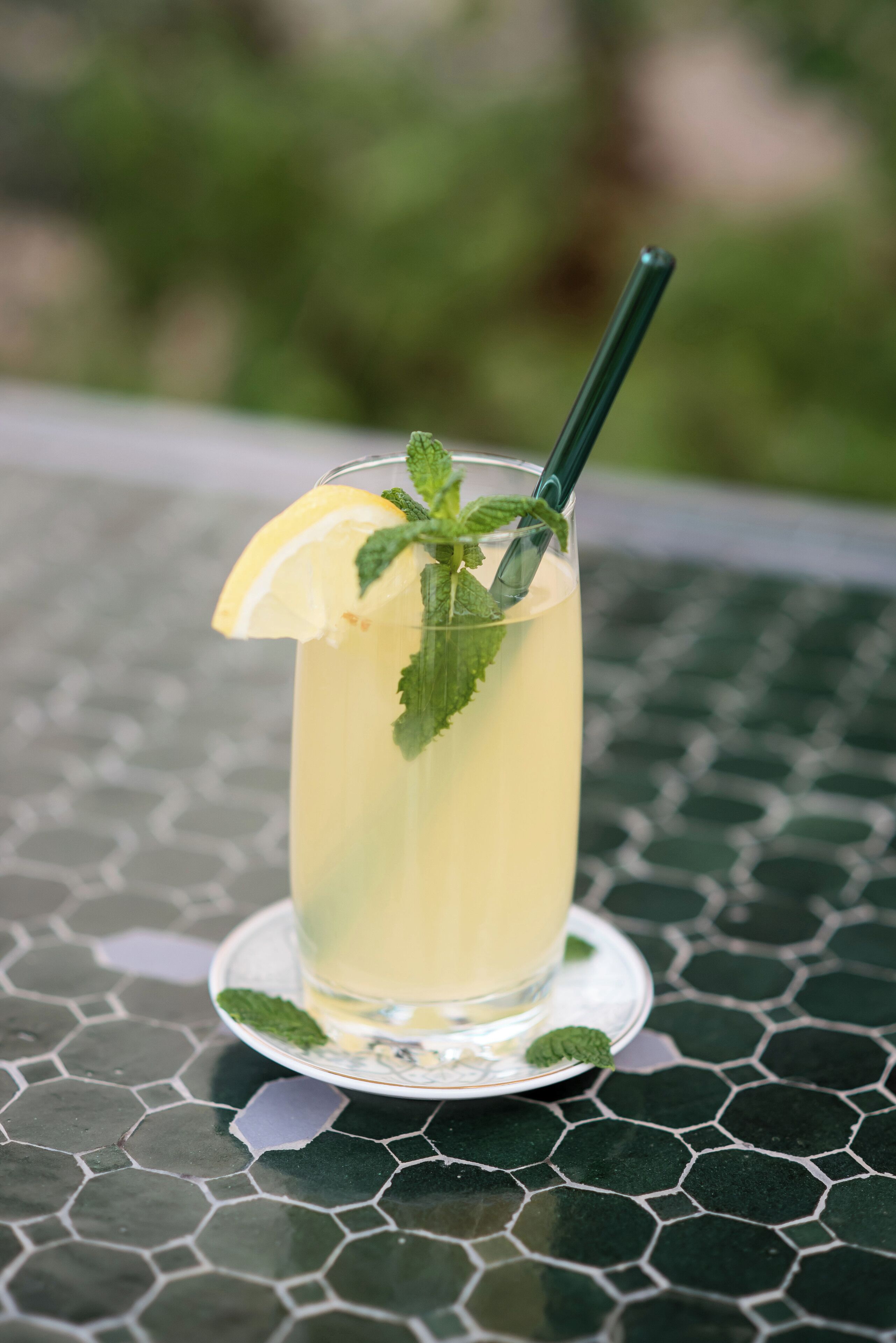 A tall glass of a yellow beverage with a lemon slice and mint garnish, placed on a patterned table.