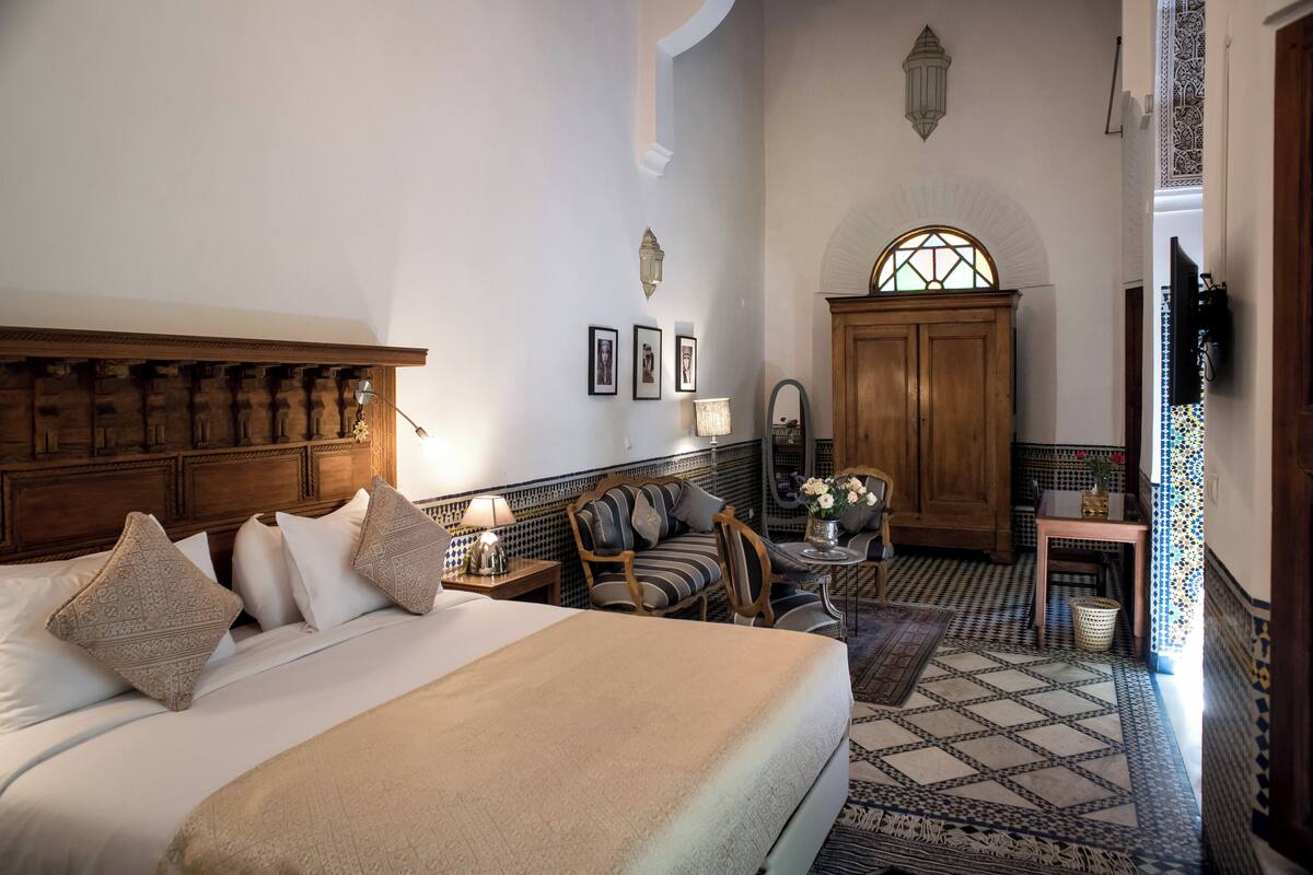 A hotel room with a large bed, a wooden headboard, a small table with a lamp, a chair, and a decorative mirror.