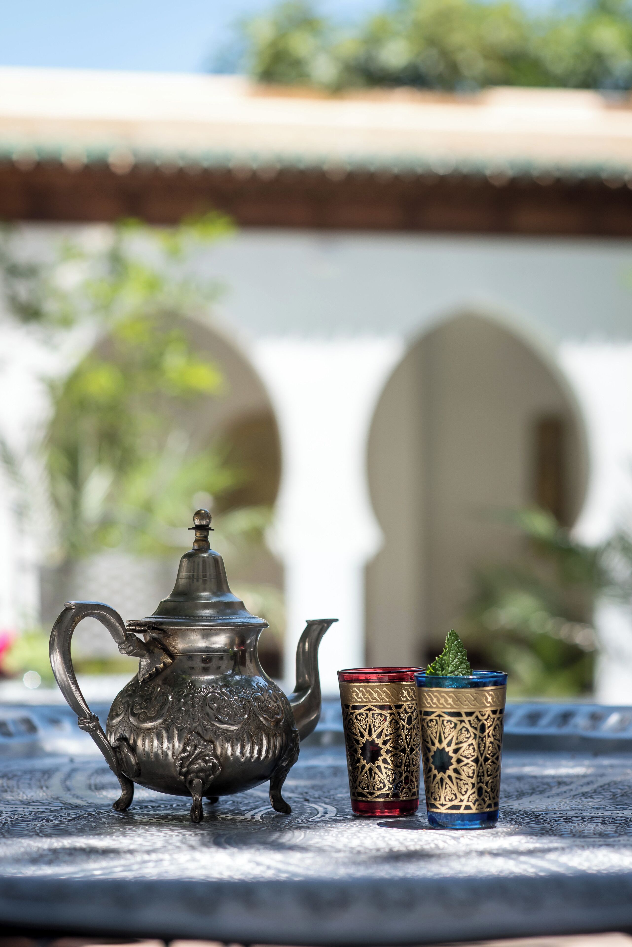 A metal teapot and two ornate glasses on a table with a tiled floor and arches in the background.
