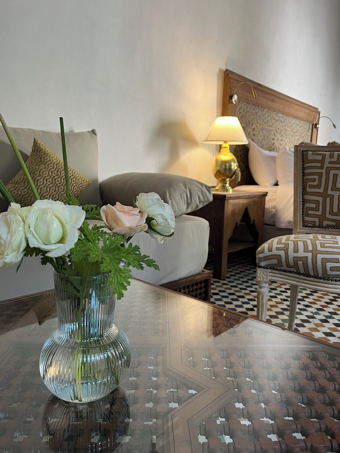 A hotel room with a bed, a nightstand with a lamp, a patterned armchair, and a table with a vase of flowers.
