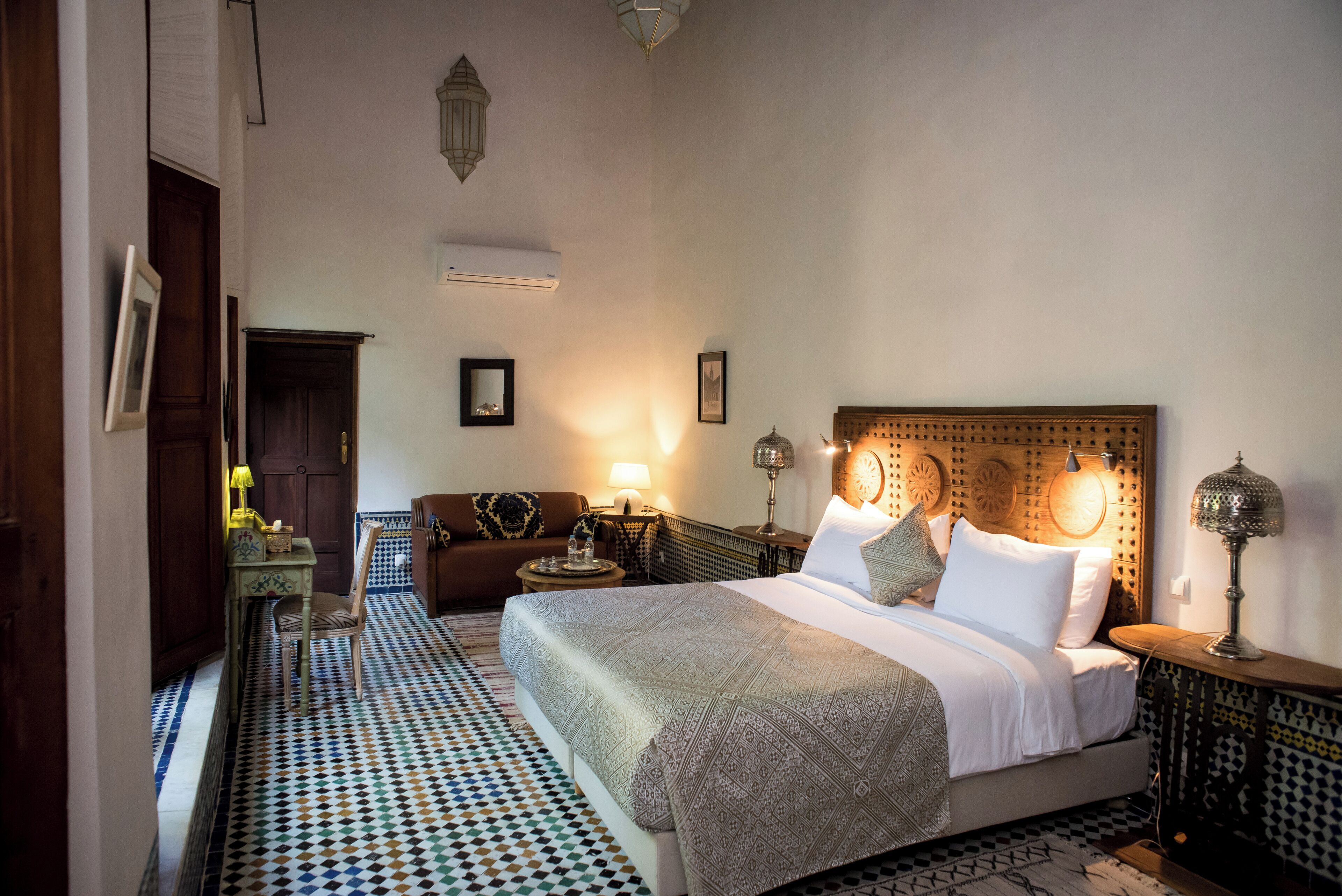 A bedroom with a large bed, a wooden headboard, a patterned floor, and a nightstand with a lamp.