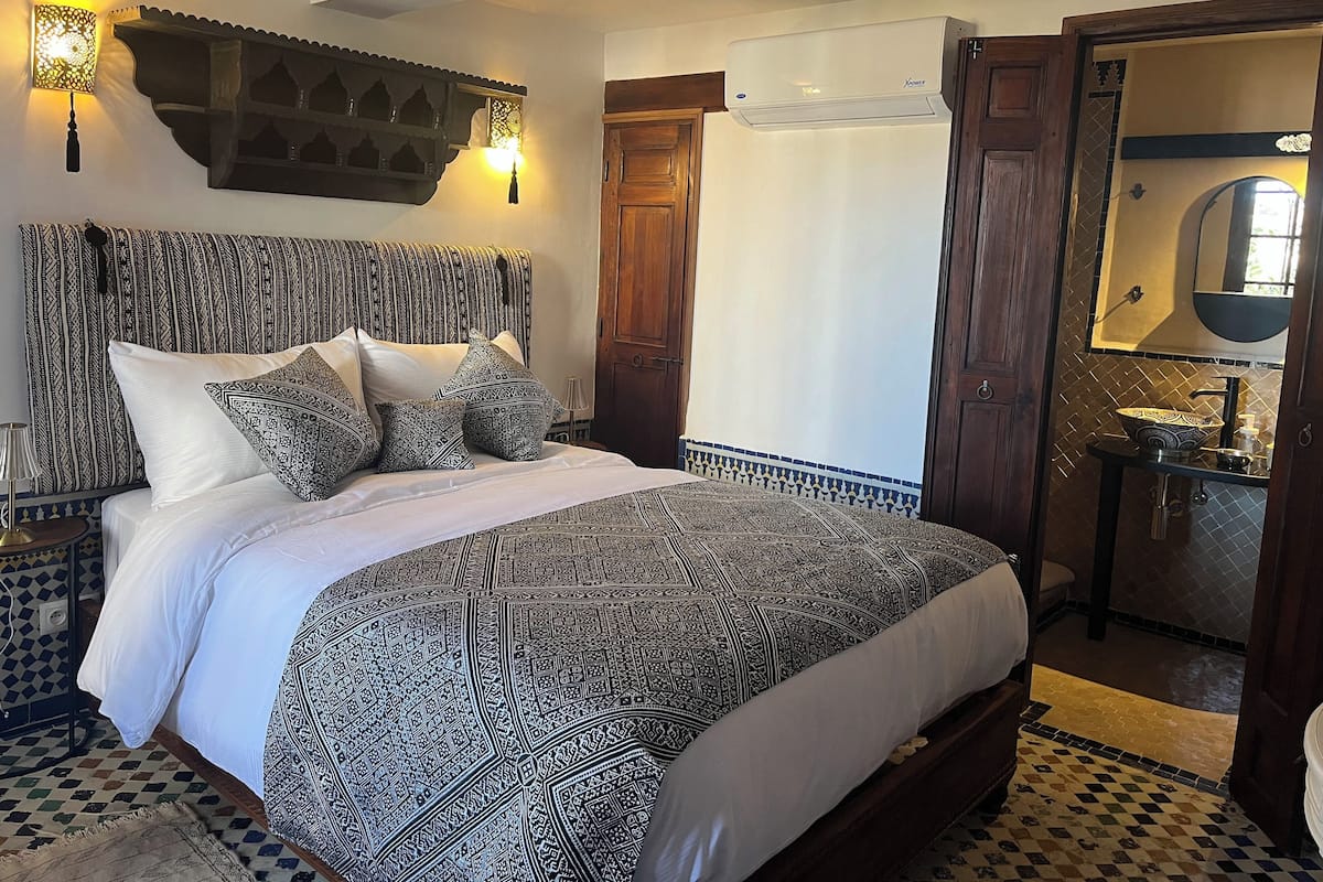 A bedroom with a patterned bedspread, a headboard with intricate designs, a wall-mounted light fixture, and a bathroom with a sink and mirror.